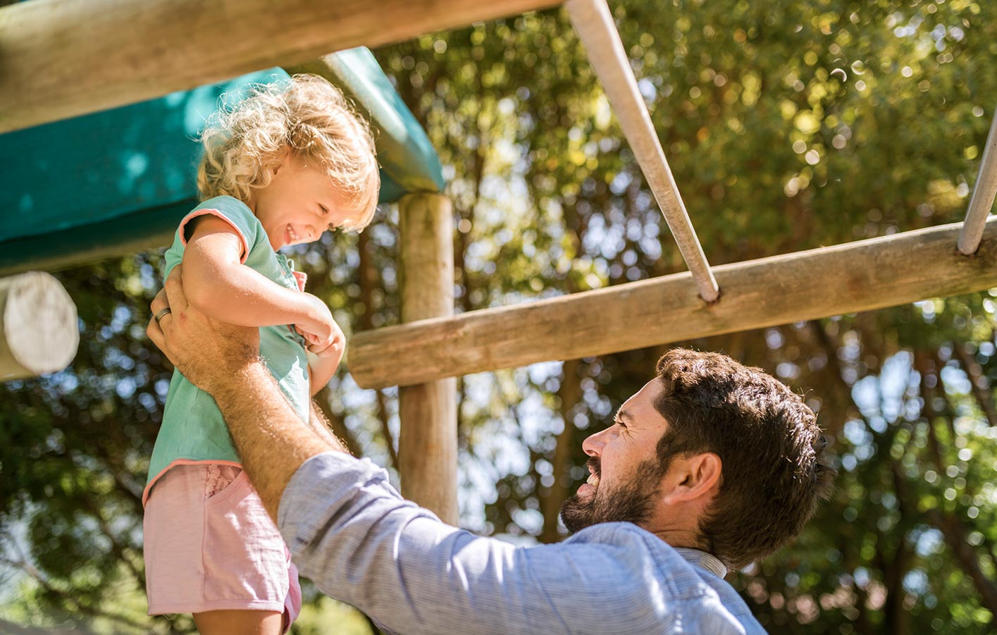 Vater spielt mit Kind am Holzspielgerät Vater hebt kleines Kind an einem Spielgerät aus Holz im Garten hoch