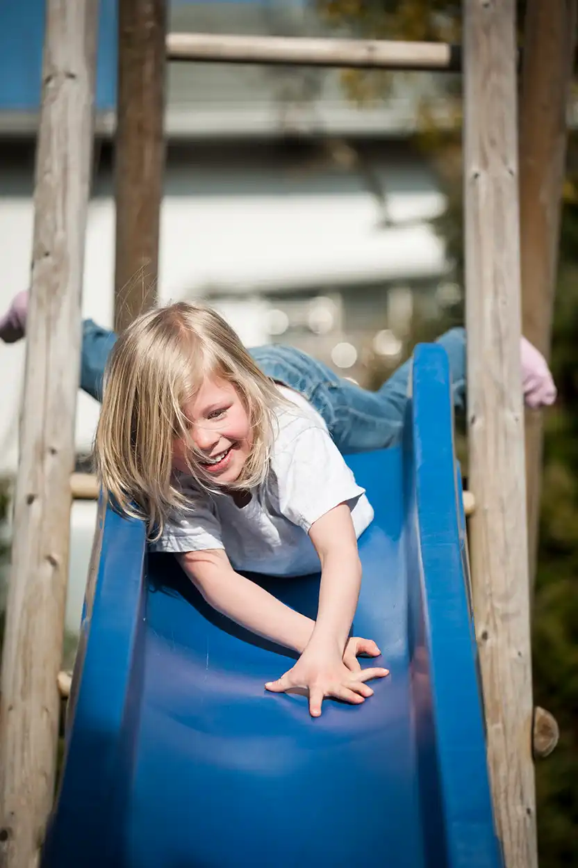 Mädchen auf Rutsche eines Spielgeräts aus Holz Ein blondhaariges Mädchen rutscht lachend auf einer blauen Rutsche eines Spielturms
